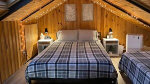 A cozy attic bedroom with a sloped ceiling, wooden walls, a central bed with a plaid bedspread, two bedside tables with lamps, and a small white heater.
