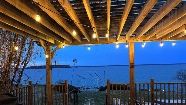 A wooden deck with string lights, a view of the water, and a metal railing. - Cozy Lake House on Oneida with Waterfront Access (Cicero)