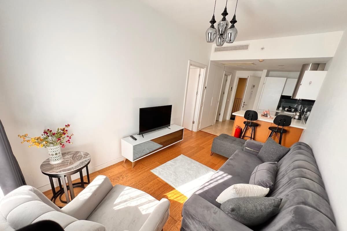 A modern living room with a grey sofa, a white armchair, a TV on a white stand, and a small table with a vase of flowers.