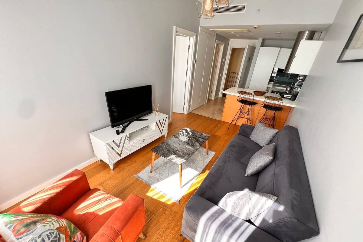 A modern living room with a flat-screen TV, two sofas, a coffee table, and a kitchen area in the background.