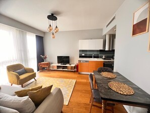 Una sala moderna con sofá, una mesa de comedor con individuales, televisor y una cocina con armarios y estufa.