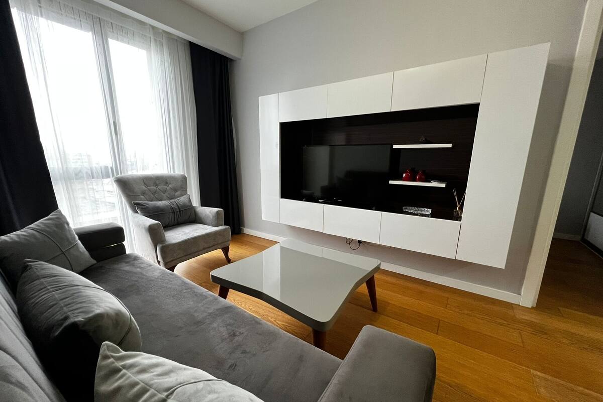 A modern living room with a large flat-screen TV mounted on a white wall, a grey sofa, a matching armchair, and a coffee table.