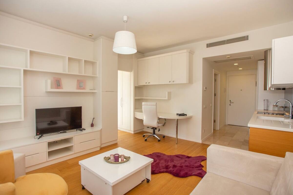 A modern living room with a TV, white furniture, a desk, and a kitchen area.