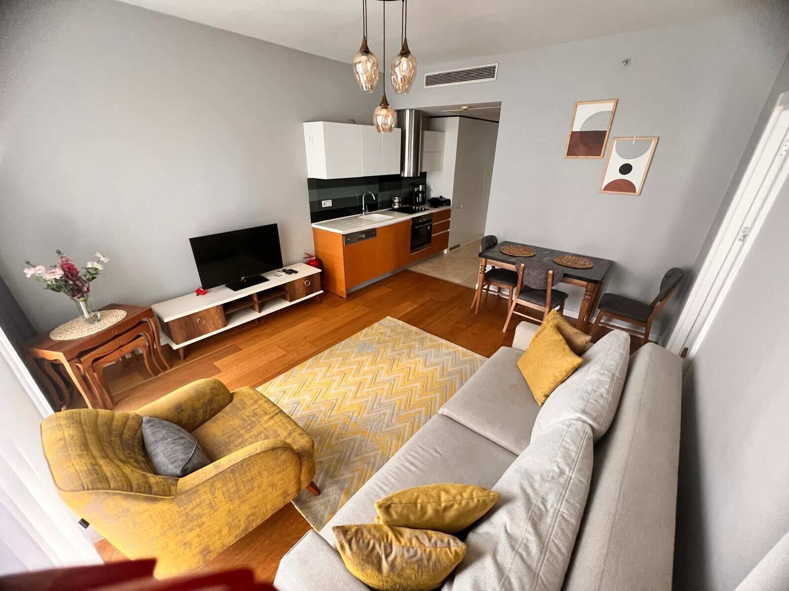 A modern living room with a sofa, armchair, and TV, a kitchen with white cabinets, and a dining area with a table and chairs.