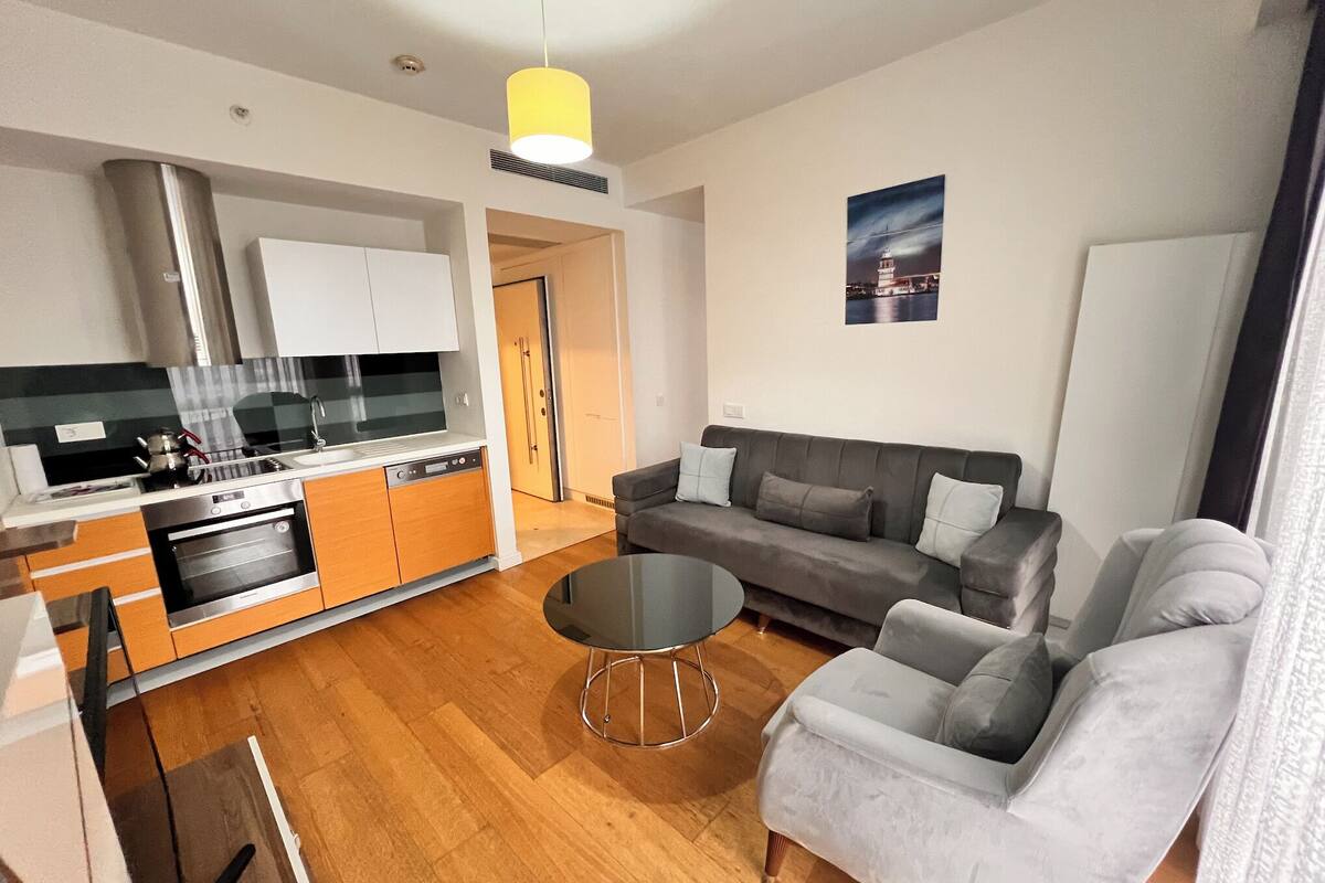 A modern living room with a grey sofa, a round coffee table, and a kitchen area with white cabinets and a stainless steel range hood.