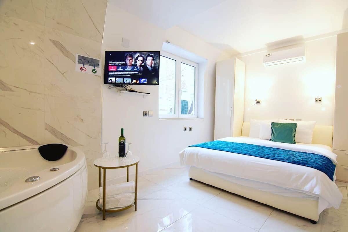 Una habitación de hotel moderna con una cama grande, un televisor montado en la pared, una mesita de noche con una botella y vasos, y una bañera.