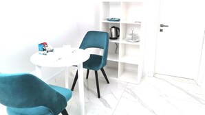 A modern room with a white door, a white round table, two teal chairs, and a white shelving unit with a kettle and a small table. - Lumiere Luxe Suites (Belgrade)