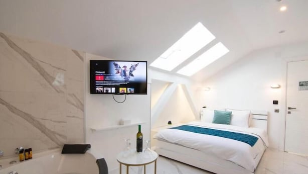 A modern hotel room with a large bed, a TV mounted on the wall, a bathtub, and a skylight. - Lumiere Luxe Suites (Belgrade)