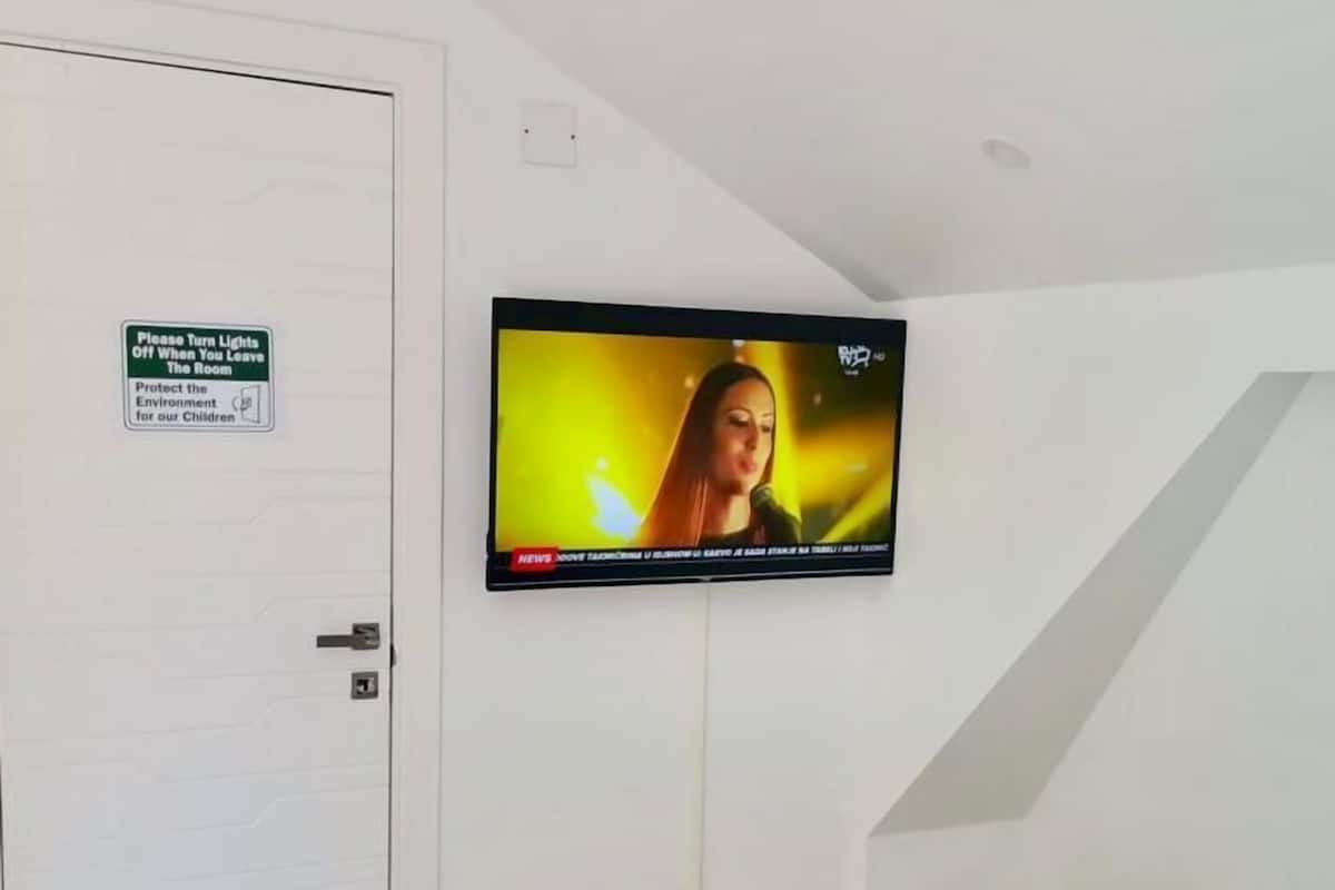 Habitación de hotel con una televisión montada en la pared, cama con detalles azules y una puerta con un letrero que indica apagar las luces para proteger el medio ambiente por los niños.