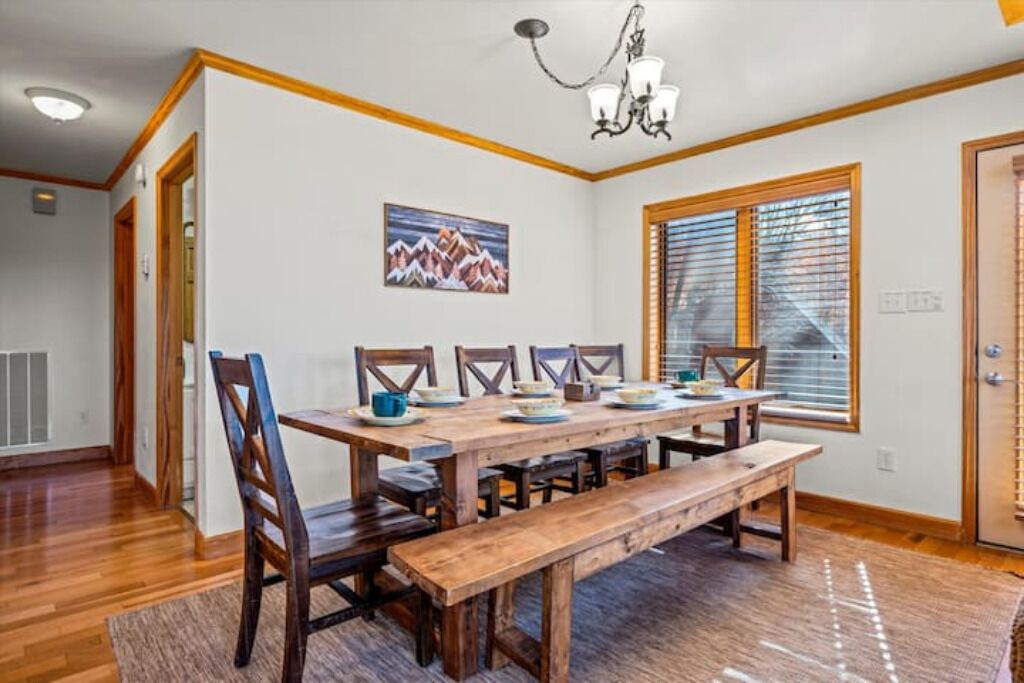 A dining area with a wooden table, chairs, and a bench. There is a chandelier above the table and a window with blinds to the right.