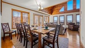 A spacious living area with a wooden dining table, chairs, and a sofa. Large windows provide natural light and views of the outside. - Long Range Views Hot Tub Private FirePit Kid Bunks (Beech Mountain)