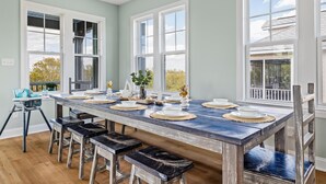 Un coin repas avec une longue table en bois, dressée pour six personnes, de grandes fenêtres et une vue sur l’extérieur.