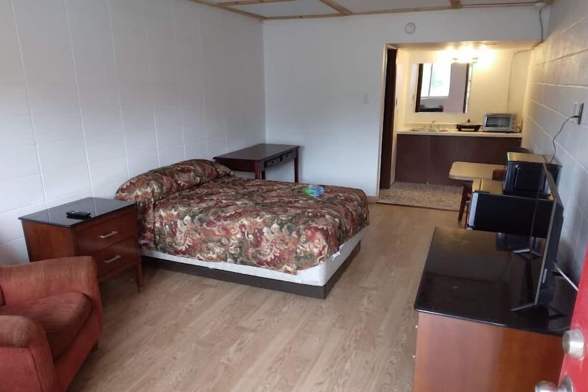 A bedroom with a bed, a small desk, a chair, a dresser, and a bathroom with a mirror and sink.