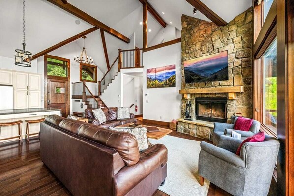 A spacious living room with a stone fireplace, leather sofa, and grey armchair. A staircase with wooden beams leads to an upper level.