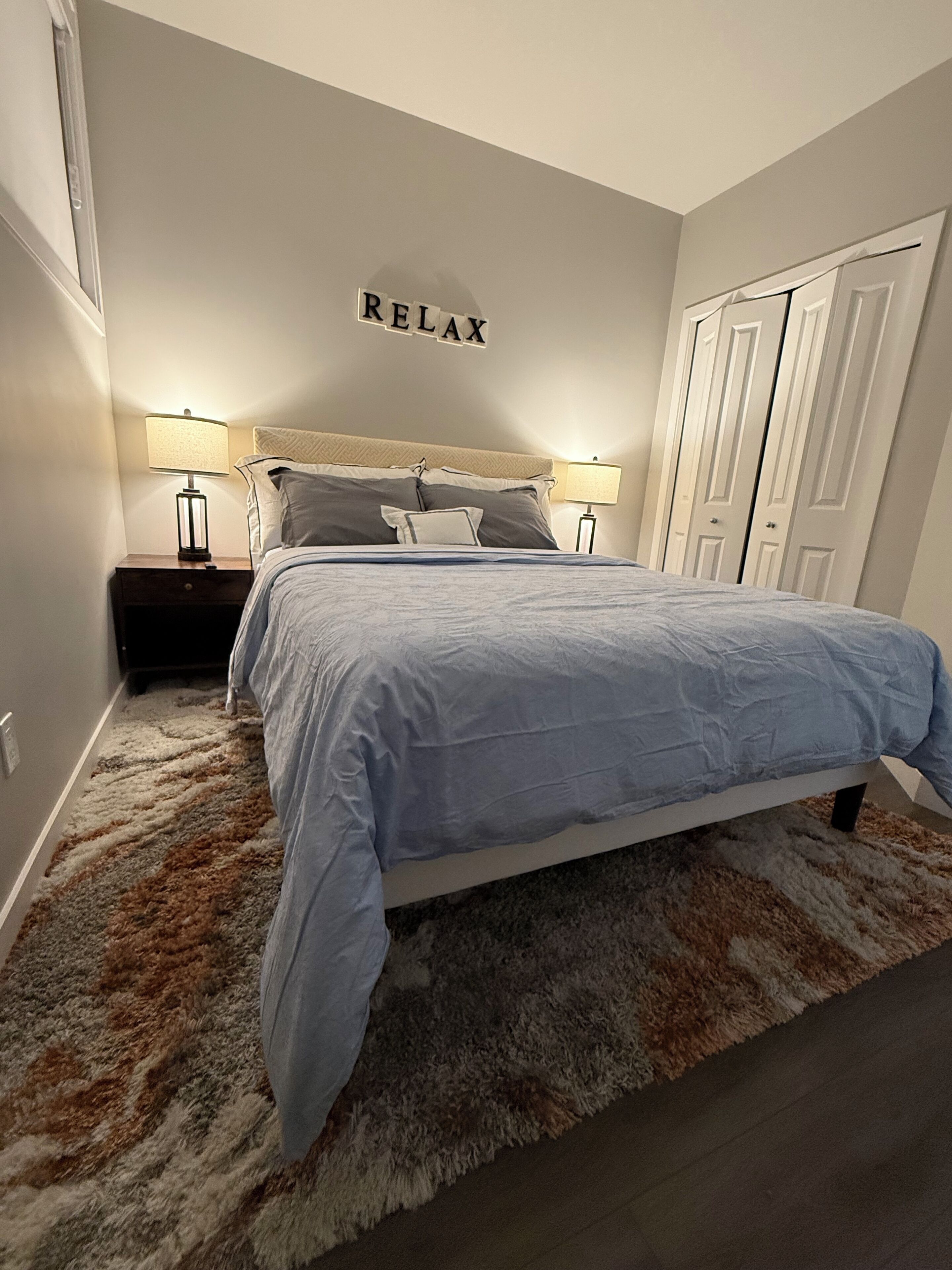 A bedroom with a bed, two bedside tables with lamps, a fluffy rug, and a wall sign that says "RELAX".