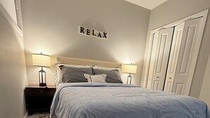 Ein Schlafzimmer mit einem Bett, zwei Nachttischen mit Lampen, einem flauschigen Teppich und einem Wandschild mit der Aufschrift 'RELAX'.