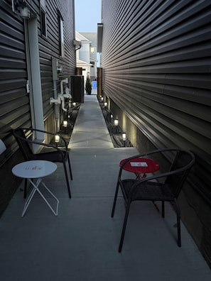 A narrow outdoor corridor with two tables and chairs, overhead lighting, and a pathway leading to a building entrance. - Almost Heaven- Near Spruce Meadows, Banff - Sleeps 8 (Calgary)