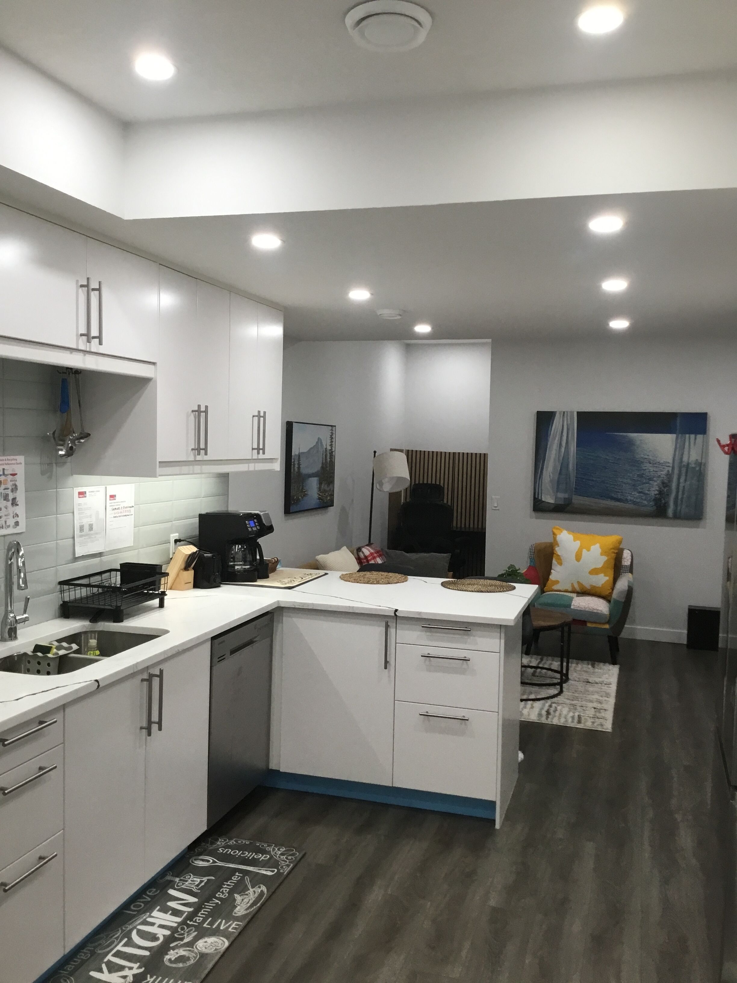 Una cocina moderna con gabinetes blancos, lavavajillas integrado y cafetera. Hay un comedor con una silla y una mesa, y una alfombra en el suelo.