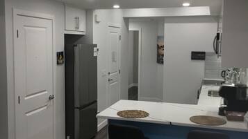 A modern kitchen with white cabinets, a stainless steel refrigerator, a bar area with stools, and a coffee machine.