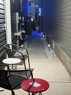 Un étroit couloir extérieur avec un chemin, deux chaises, une petite table et une boîte aux lettres.