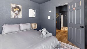Un dormitorio con una cama, almohadas, una mesita de noche, una pared decorada con obras de arte y una puerta que da a otra habitación.