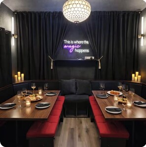 A restaurant interior with a neon sign, dark curtains, wooden tables, red cushioned benches, and candlelit ambiance.