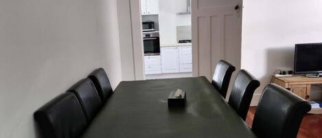A dining area with a table and chairs, a television, and a kitchen in the background.
