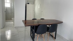 Un comedor con mesa de madera y sillas negras, suelo de baldosas blancas y pared blanca.