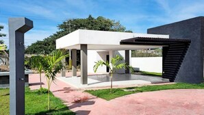 Una casa moderna con un patio cubierto, estructura de concreto y un pequeño jardín con palmeras.
