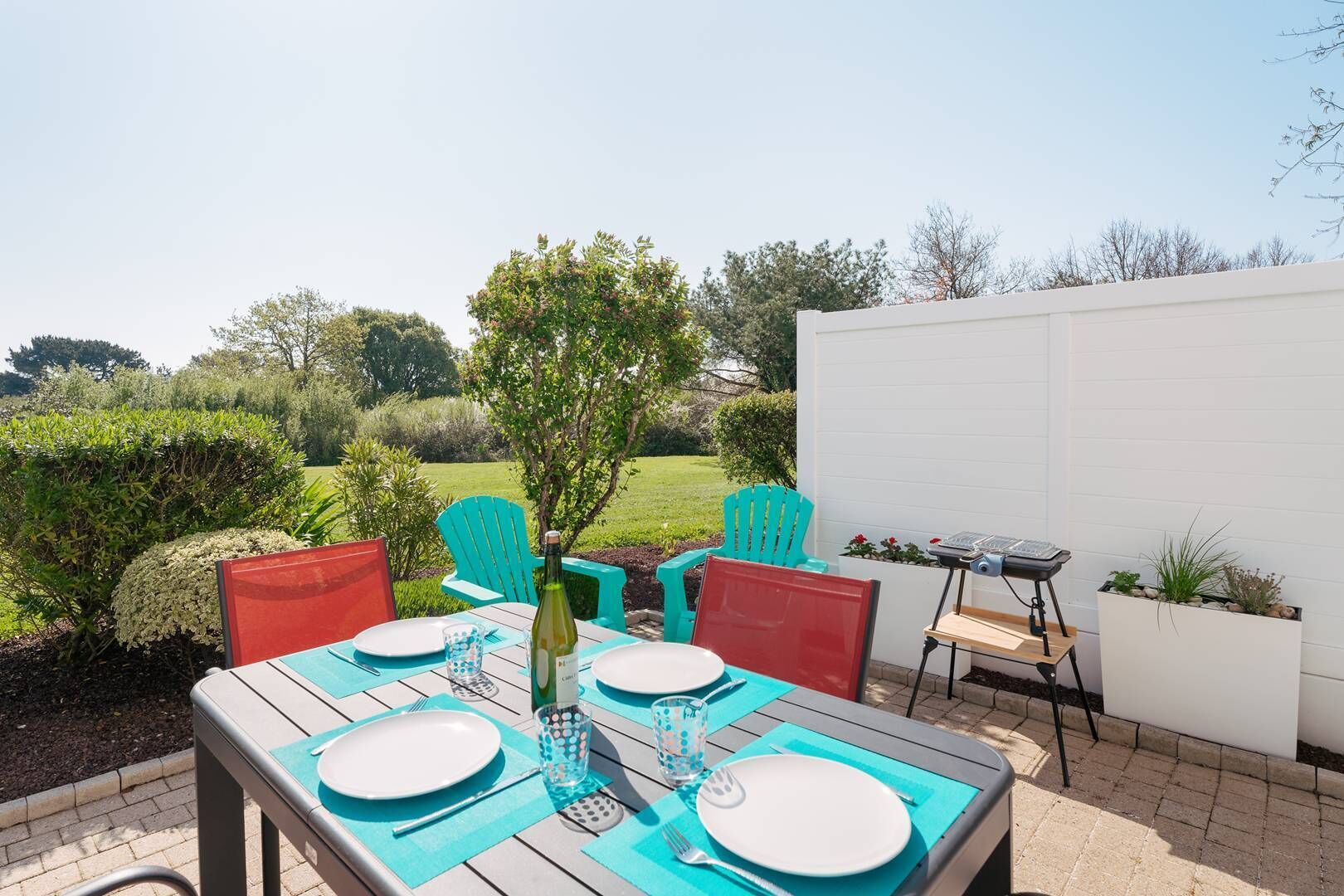 Un patio exterior con una mesa preparada para cuatro personas, rodeado de sillas, una parrilla y una pared blanca.