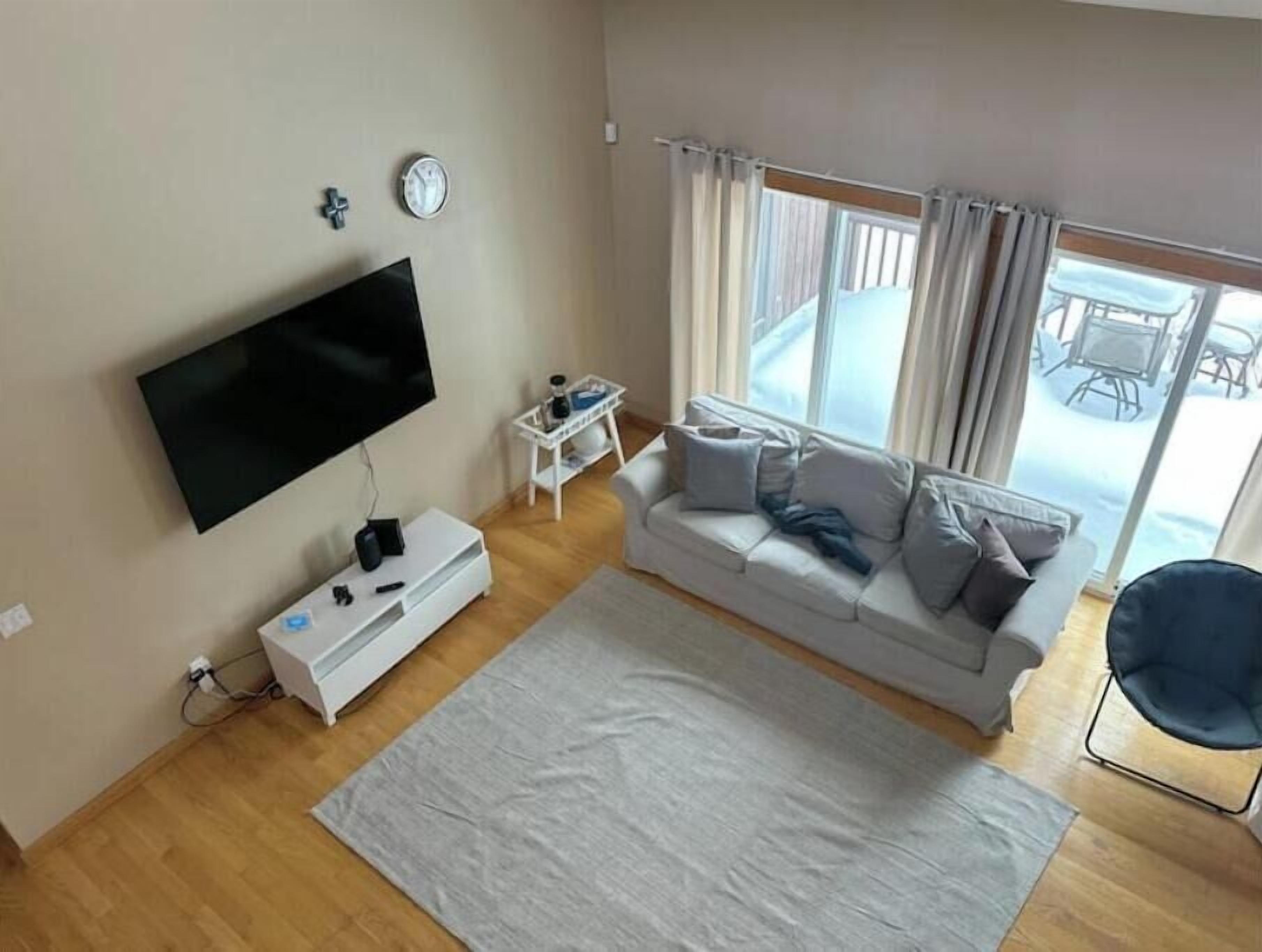 A living room with a flat-screen TV, a white sofa, a small white side table, and a large window with curtains.