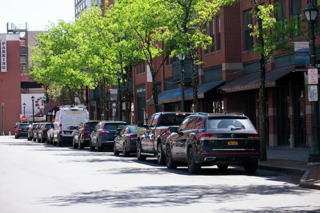 Une rangée de voitures garées le long d’une rue bordée d’arbres, avec des immeubles de chaque côté.