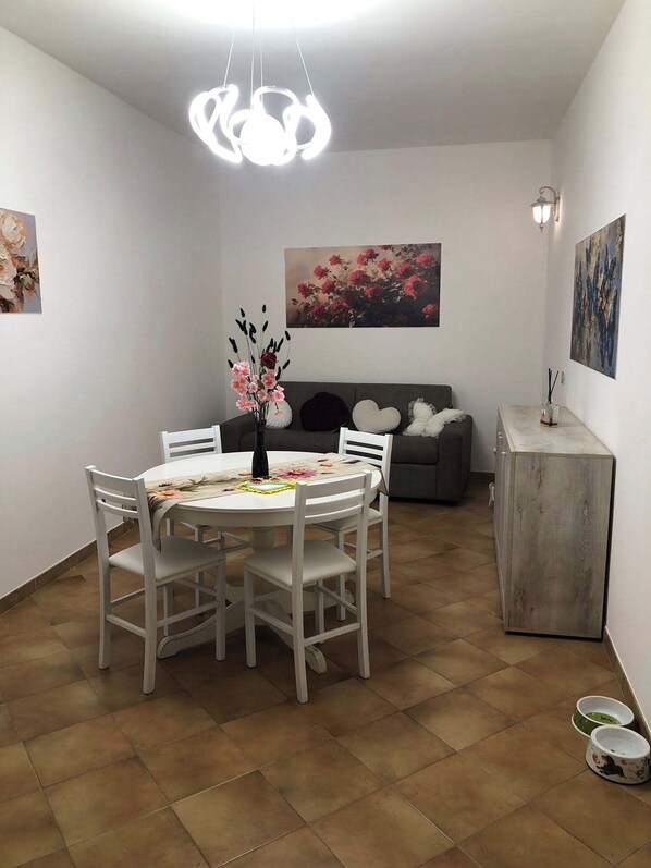 A dining area with a round table, white chairs, a vase with flowers, a sofa, and a cabinet.