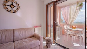Un salon moderne avec un canapé beige, une horloge en bois accrochée au mur et un balcon doté d’une table, de chaises et offrant une vue sur le ciel.