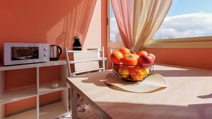 Une table près d’une fenêtre, avec des rideaux, est dressée pour le petit-déjeuner : on y trouve un four à micro-ondes, une machine à café et un panier de fruits.