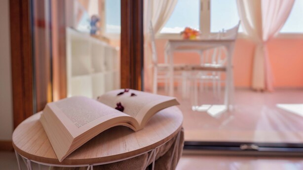 Un livre ouvert est posé sur une table ronde en bois, dans une pièce donnant sur un balcon et où se trouve une chaise blanche.