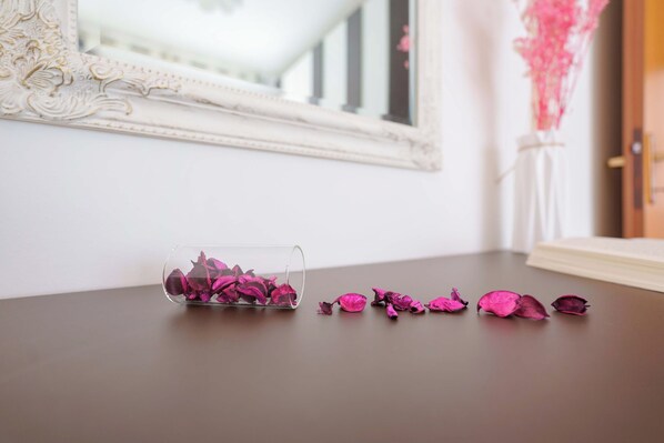 A clear glass vase with pink dried flowers on a brown table, next to a white ornate mirror and a pink decorative piece.
