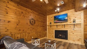 A cozy wooden cabin interior with a leather sofa, two small tables, a mounted TV, and a fireplace. - PRIVATE! 2 Miles To PF! Charming 4BR Farmhouse w/Hot Tub, PT, Fire Pit & VIEWS! (Sevierville)