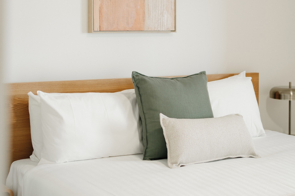 Una cama bien hecha con sábanas blancas y dos almohadas, una verde y otra blanca, en una habitación con cabecera de madera.