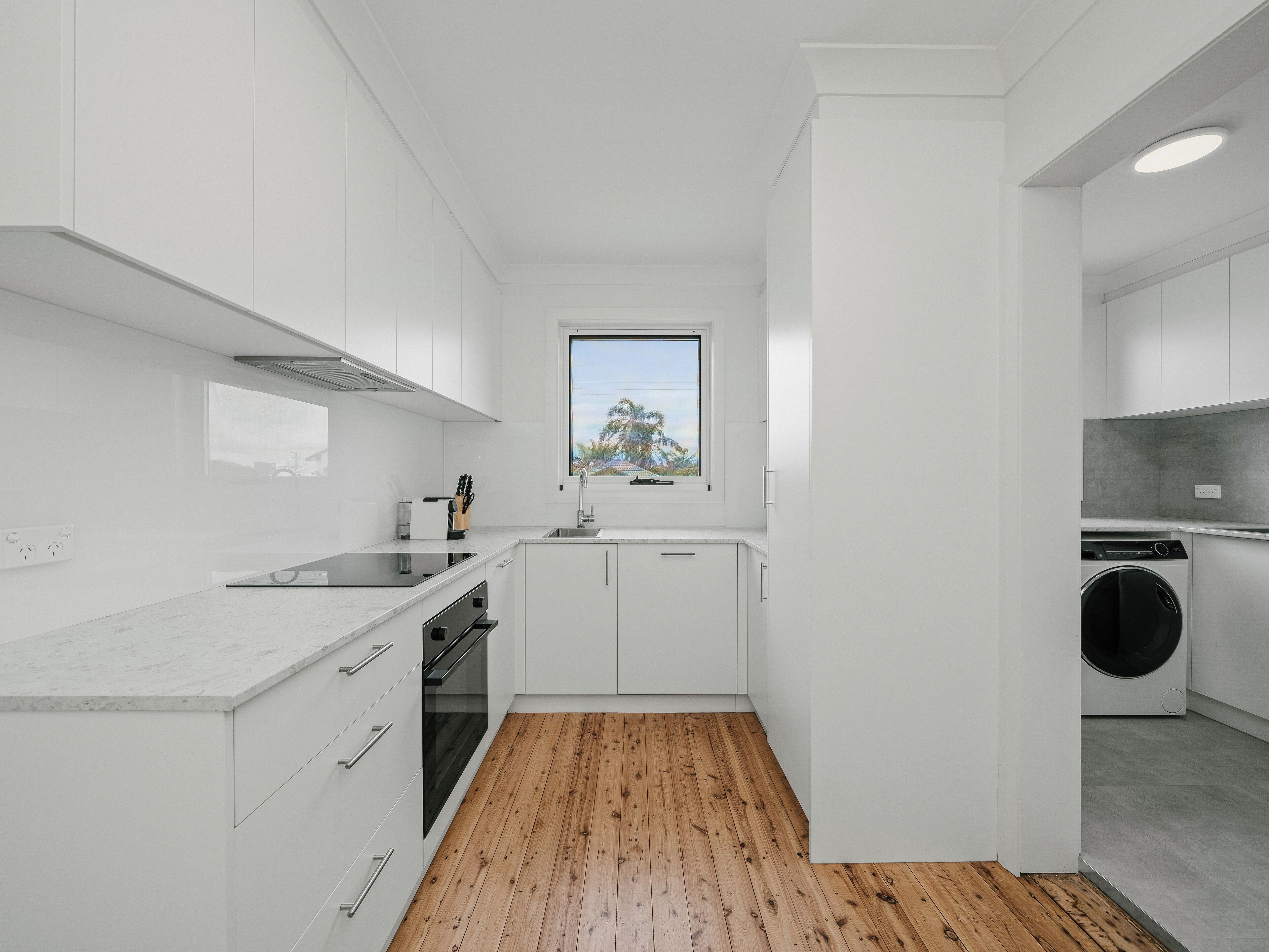 Una cocina moderna con armarios blancos, suelo de madera, horno empotrado y ventana con vistas a palmeras.