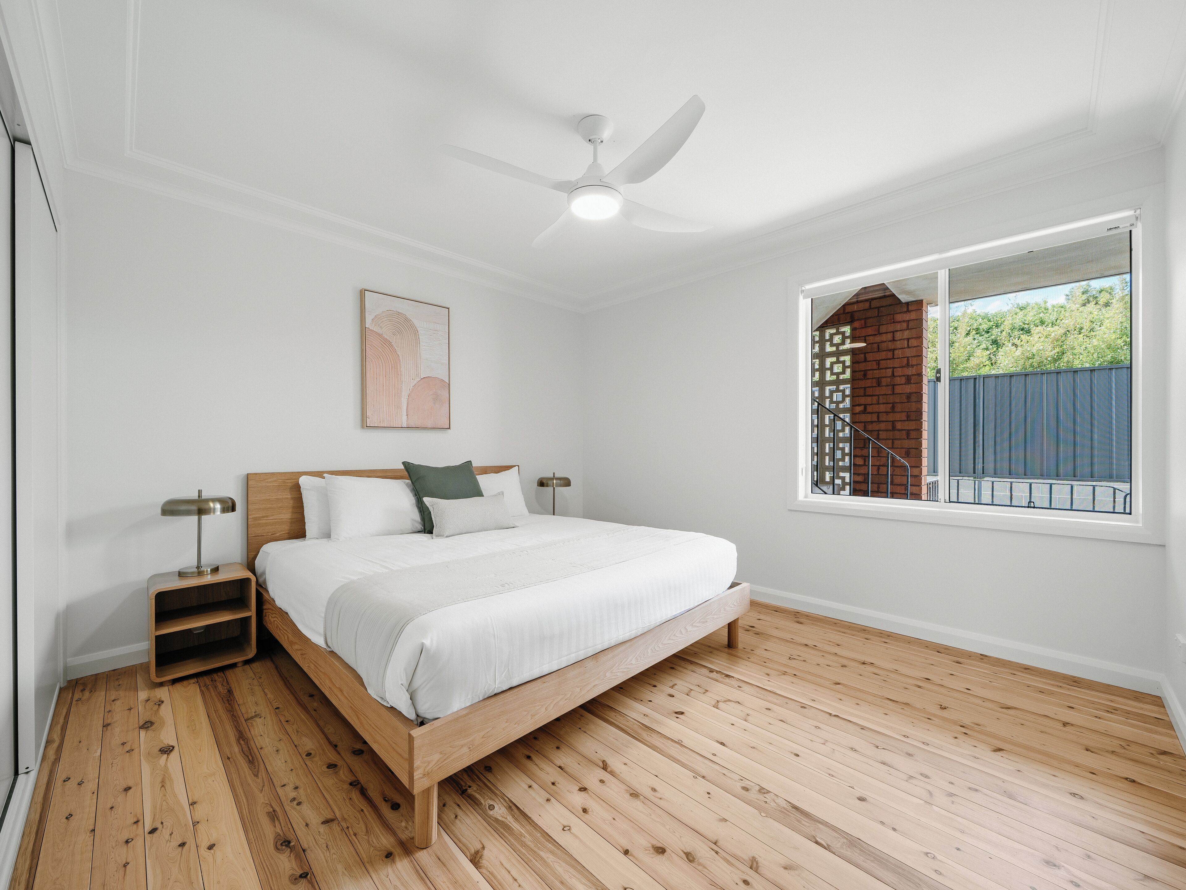 Un dormitorio con cama de madera, paredes blancas, ventilador de techo, ventana con vistas a un muro de ladrillos y follaje, y una mesita de noche con lámpara.