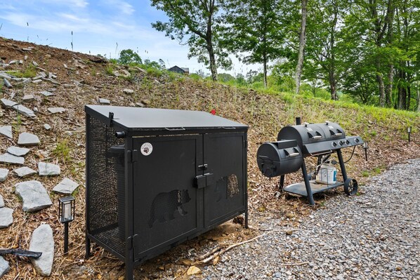 Een draagbare barbecue en een metalen opbergkist met een berenklauw logo, staan op een grindpad met bomen op de achtergrond.