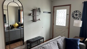 A living room with a grey sofa, a bench, a mirror, a wall clock, and a door with a stained glass window. - Cozy 3-bedroom house with a pool. Close to SPAC, Track and Downtown (Ballston Spa)
