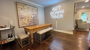 A modern living room with a wooden bench, a piano, a large abstract painting, and a neon sign that reads "GET DOWN TIGER". - Stylish 3BR in Garden District near parks, river views, Neville High, and ULM. (Monroe)