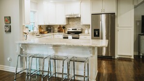 A modern kitchen with white cabinets, a stainless steel refrigerator, a built-in oven, and a marble island with bar stools. - Stylish 3BR in Garden District near parks, river views, Neville High, and ULM. (Monroe)