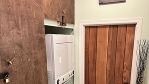 A laundry room with a washer and dryer, a wooden door, and a landscape painting on the wall. - Island paradise with private hot tub -- pet friendly & breakfast included! (Pender Island)