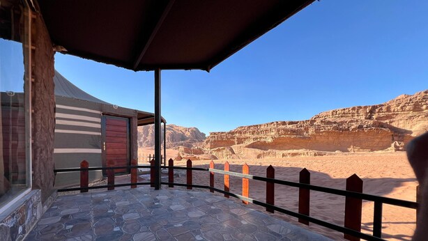 A stone-tiled balcony with a view of a desert landscape, a wooden fence, and a building with a striped awning. - Room-wadi RUM Guide, Camp & Tour (Wadi Rum)