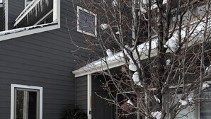 A two-story house with a dark exterior, a front door, and a small porch area with blue Adirondack chairs.