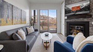A modern living room with a grey sofa, a blue armchair, a white coffee table, and a fireplace. There is a large painting on the wall and a TV mounted above the fireplace. - Luxury 1BR Condo at The Mason, AC, Jordanelle, Pool & Hot Tub Access (Heber City)