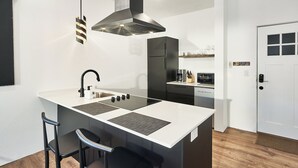 A compact kitchen with a central island, a built-in oven, and a microwave. There are two bar stools and a black chair. A white door with a 'Please remove your shoes' sign is visible. - Penthouse studio suite, downtown White Salmon, suite 15 (White Salmon)